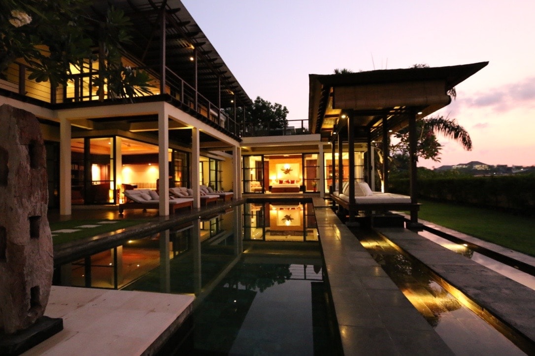 A modern villa is showcased, featuring a tranquil pool that reflects the evening sky. Large windows illuminate the interior, while outdoor lounging areas are situated under a traditional roof. Lush greenery surrounds the property, enhancing the serene environment.