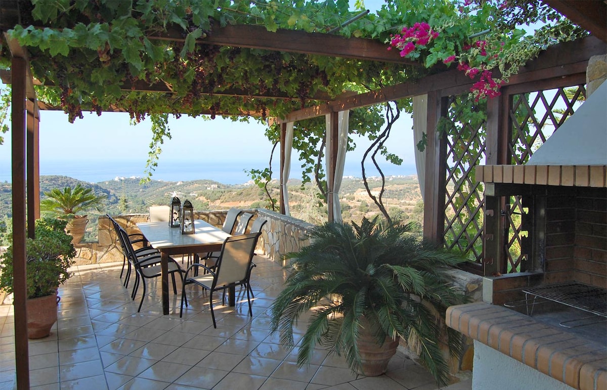 An outdoor dining area is shaded by a wooden pergola adorned with climbing vines and flowers. A long table is surrounded by several chairs, with a stone barbecue nearby, and a view of the distant sea visible in the background.