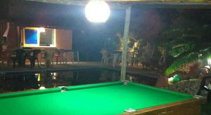 A well-lit outdoor area features a green billiard table in the foreground. In the background, a shimmering pool reflects surrounding lights. Chairs can be seen arranged around a space that indicates a relaxed social environment, enhanced by the presence of palm leaves framing the scene.