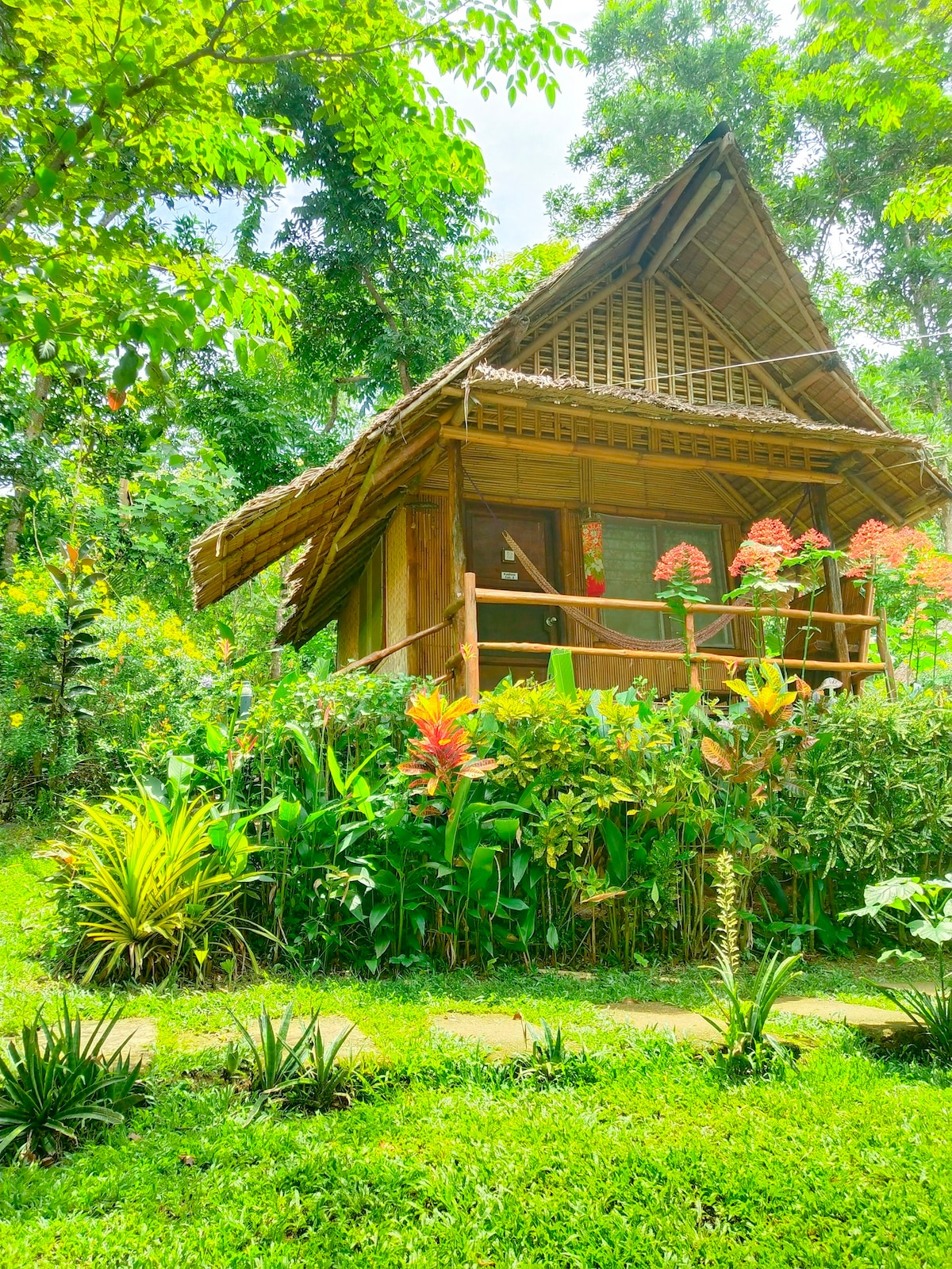 The native cottage is framed by lush greenery and vibrant flower gardens. The structure features a thatched roof and wooden walls, with large windows that provide views of the surrounding nature. Pathways lined with plants lead to the entrance, enhancing the serene environment.