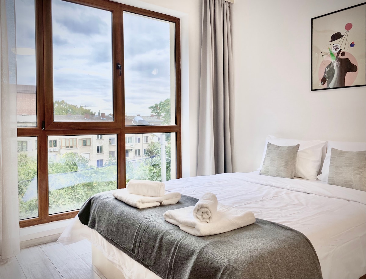 A bright sleeping area features a spacious bed, adorned with soft linens and two neatly rolled towels. Large windows allow natural light to fill the room, offering views of greenery outside. A piece of artwork hangs on the wall, adding a touch of color.