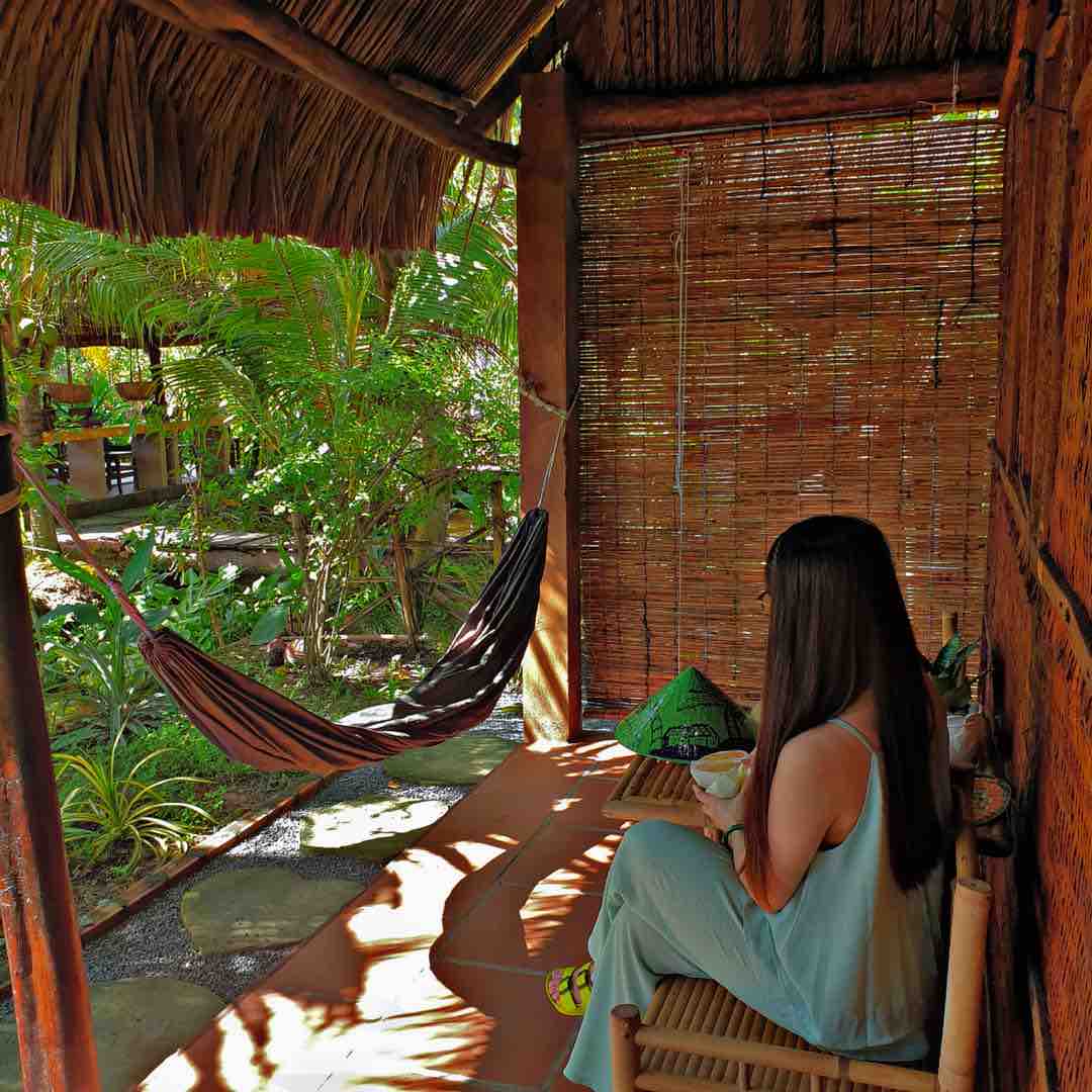 A shaded terrace features a hammock and natural decor, surrounded by lush greenery. A person is seated, enjoying a meal, with a view of tropical plants creating a serene outdoor space. Natural textures and warm tones are present throughout the area.
