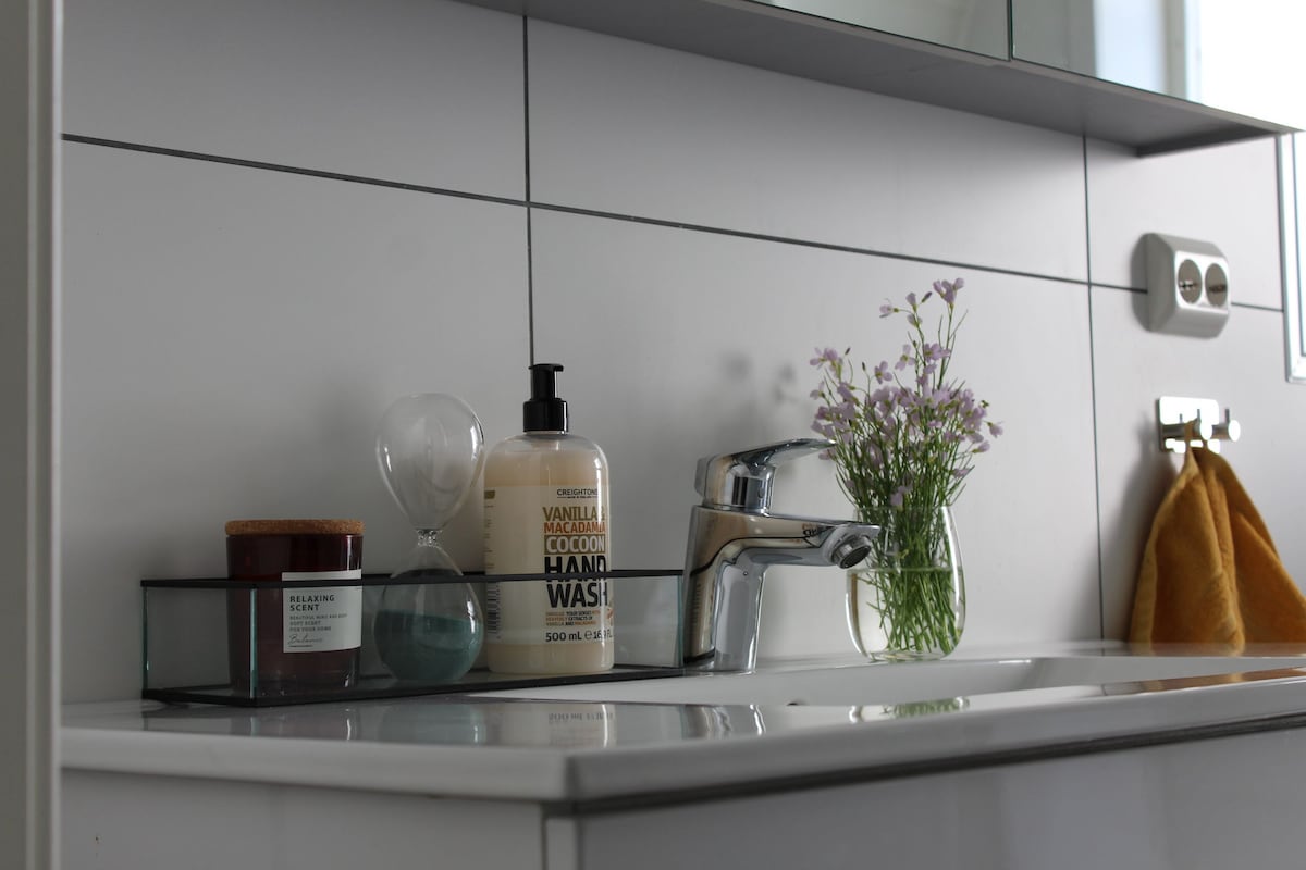A modern bathroom sink area is presented, featuring a sleek faucet and neatly arranged toiletries. A clear container holds hand wash and a candle, while a small vase with flowers adds a touch of nature. A soft towel hangs nearby for convenience.