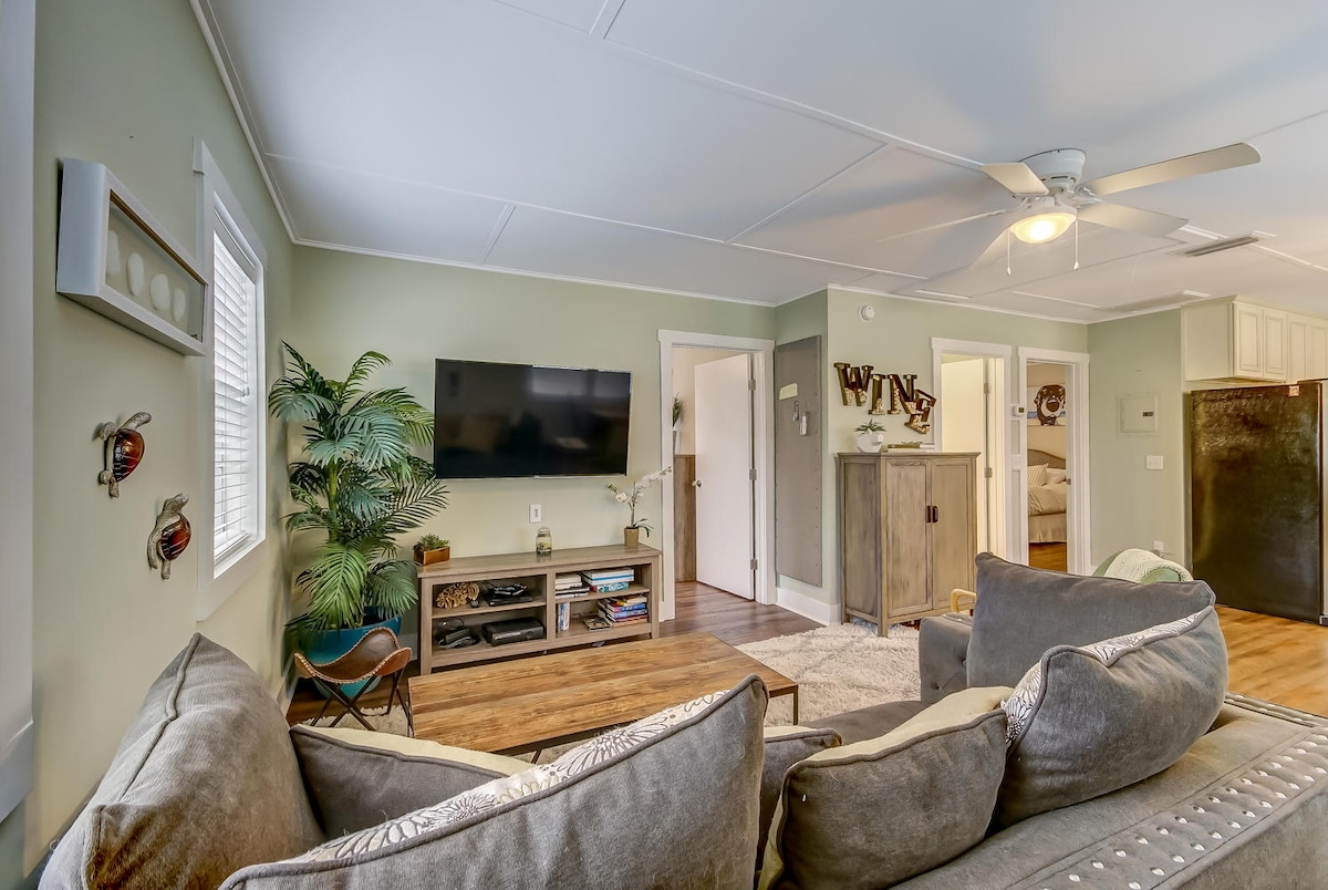 A living area is presented with a comfortable sectional sofa, accentuated by decorative pillows. A flat-screen television is mounted on the wall opposite. Light green walls create a calming environment, while a ceiling fan adds functionality to the space. A door leads to other rooms, enhancing accessibility.