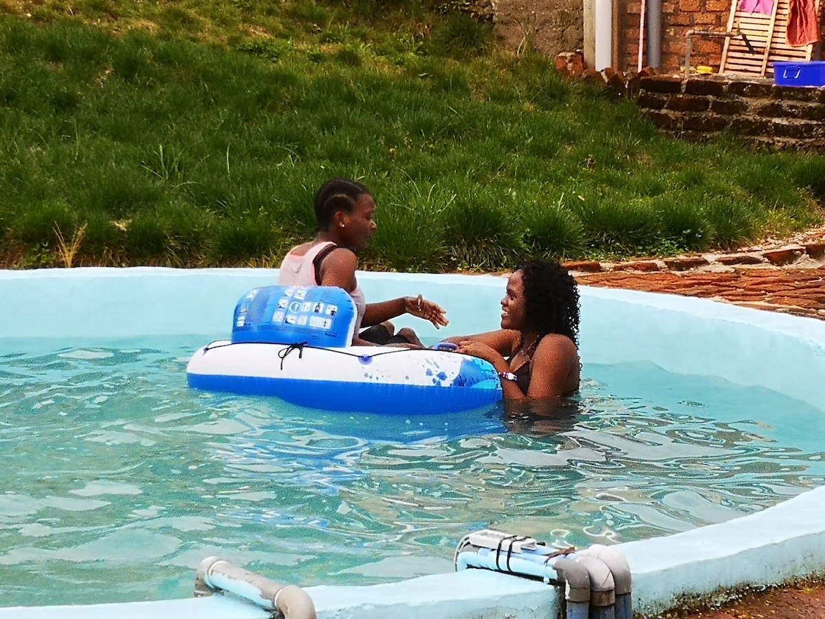 A circular pool is depicted with clear blue water, featuring two individuals enjoying their time on a floating device. Lush green grass surrounds the pool area, creating a relaxing outdoor space. Stone paths are visible in the background, enhancing the setting.