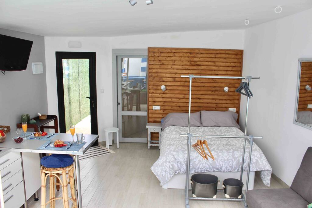 Luminoso estudio con cama de matrimonio de 135 cm en tonos grises, cuatro almohadas y un cabecero de madera barnizado. Lámparas individuales a cada lado y una mesilla de noche em el lado izquierdo.