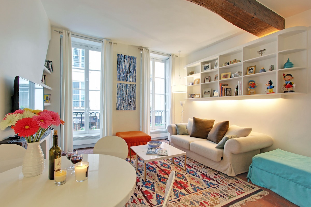 The living room is bright and spacious, featuring two large windows that let in natural light. A cozy sofa and an ottoman accompany a round dining table set for four. Decorative shelves display various items, adding character to the space.