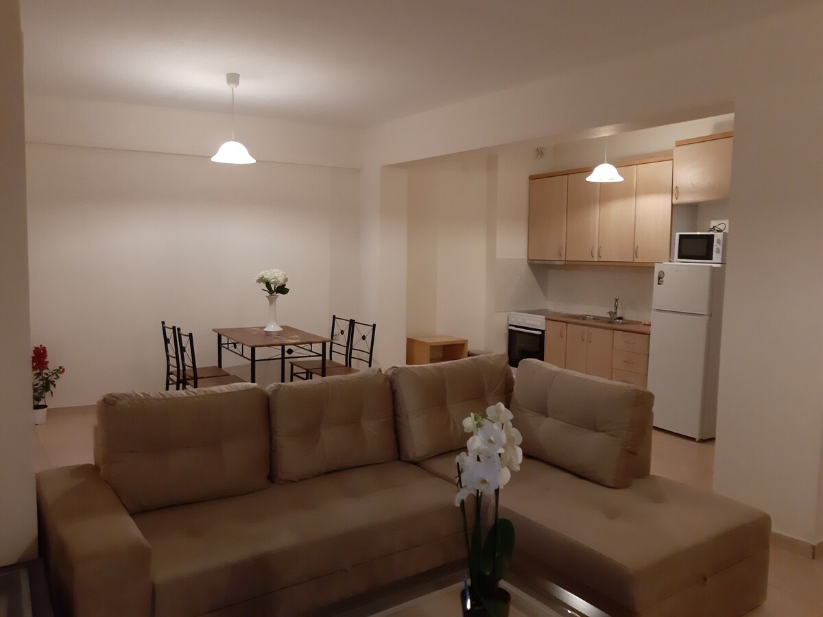 The living room features a spacious, neutral-colored couch facing a dining area with a rectangular table and six black chairs. A white flower arrangement decorates the coffee table. In the background, the kitchen is visible, equipped with modern appliances, including a refrigerator and oven.