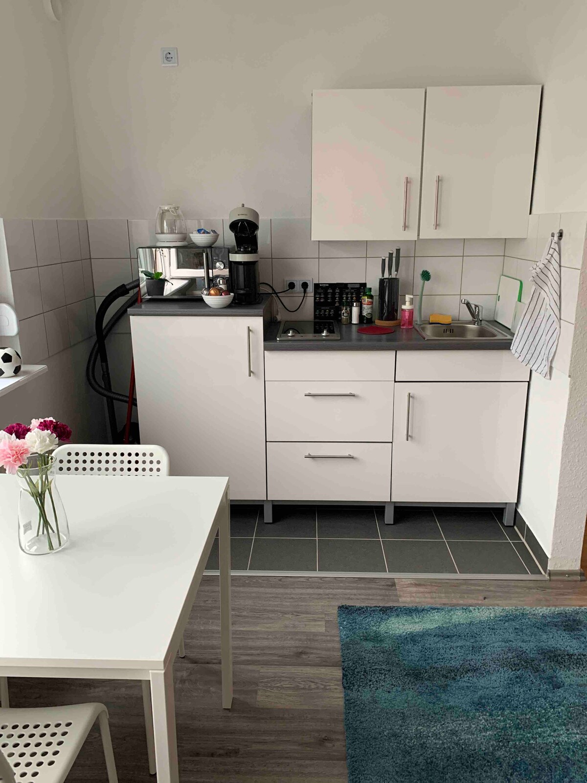 A compact kitchen area is shown, featuring modern white cabinets and a black countertop. Kitchen appliances, including a coffee machine, are visible. A small dining table with white chairs and a vase of flowers is included, while a rug adds a touch of color to the space.