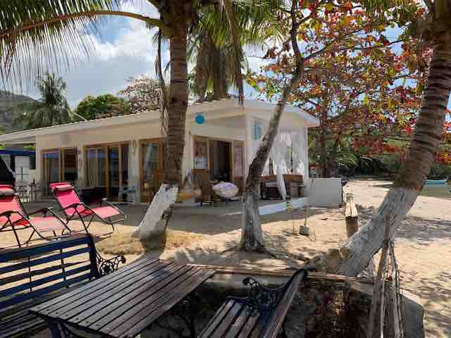 A beach front villa is situated under swaying palm trees, featuring spacious outdoor seating and tables. The entrance to the villa is visible, with a backdrop of sandy beach and vibrant foliage. Sun loungers are arranged nearby for relaxation.