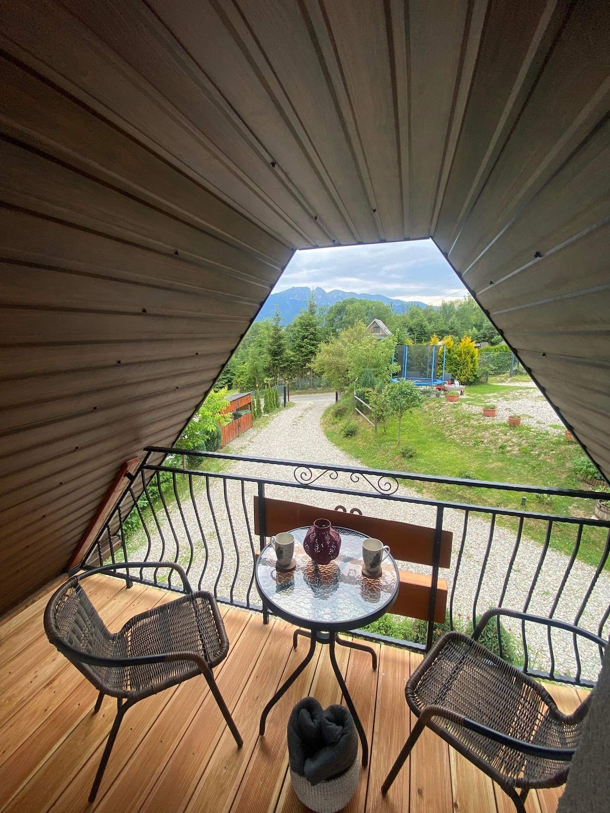 A balcony space features two woven chairs and a small round table. A scenic view of greenery and distant mountains is framed by the balcony's wooden structure. The flooring is made of warm wood, creating an inviting outdoor atmosphere.