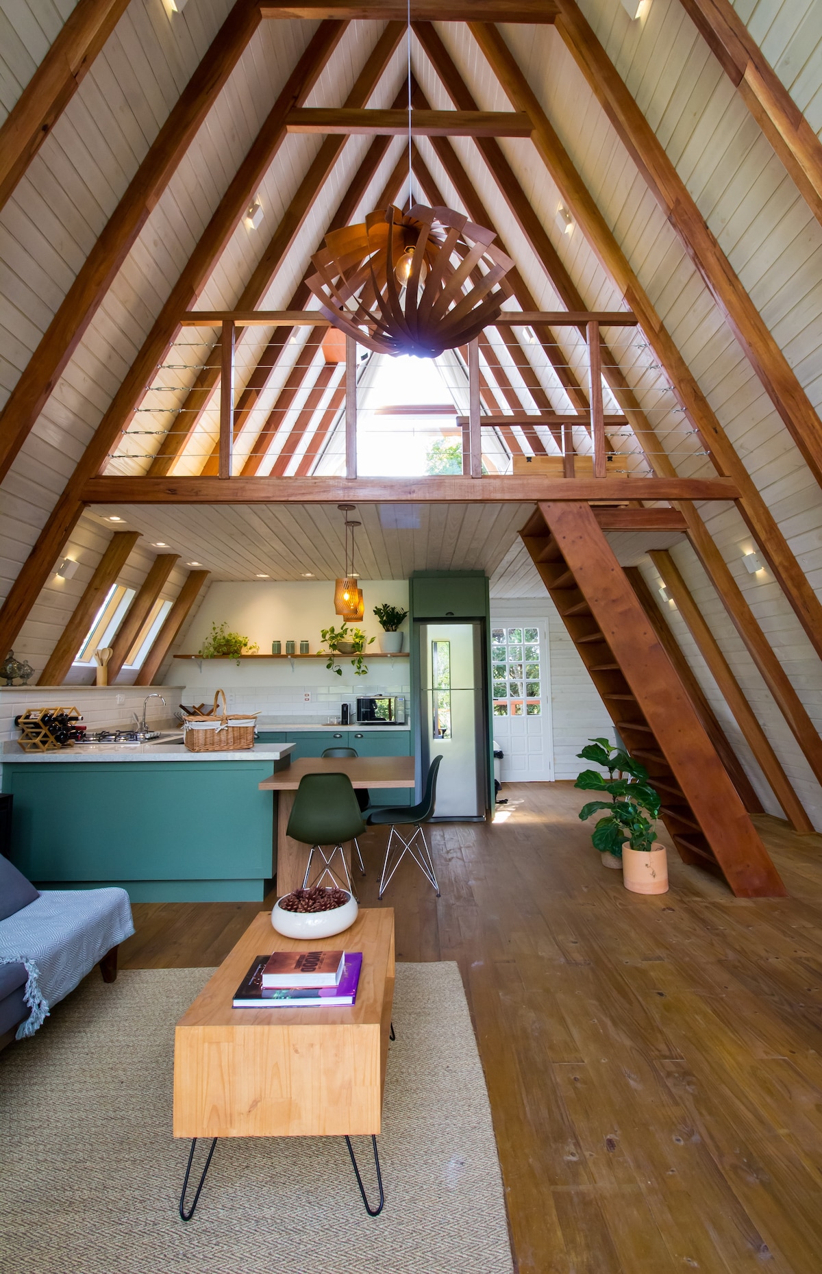 The interior of the A-Frame cabana features high, wooden ceilings with exposed beams, creating an airy atmosphere. A spacious living area includes a comfortable sofa and a wooden coffee table. The open kitchen is visible, equipped with modern appliances, and natural light floods the space through large windows.