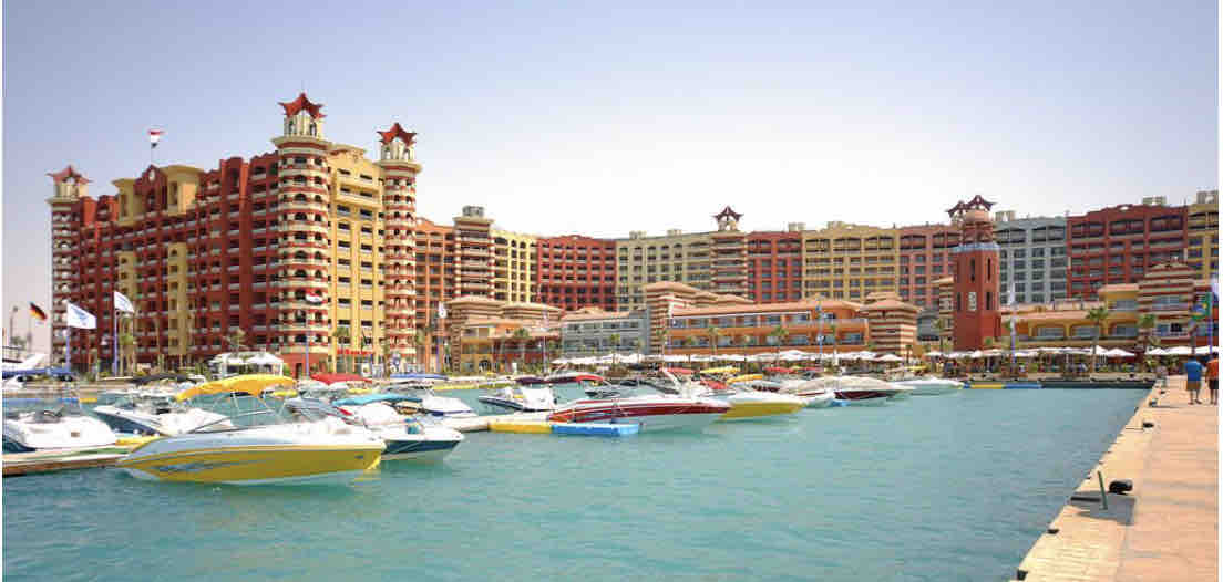 A waterfront view showcases an array of colorful boats anchored on clear blue water. Towering buildings with distinctive architectural features line the shore, adorned with star motifs. The atmosphere reflects a vibrant marina setting, ideal for leisurely strolls along the promenade.