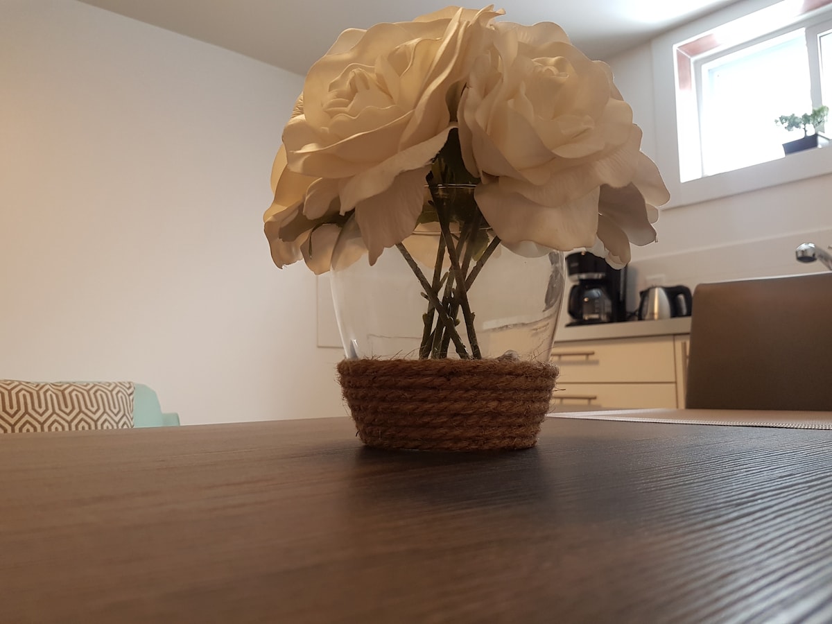 A glass vase filled with white flowers is positioned on a wooden table. The vase is wrapped with a natural jute twine. Minimalistic kitchen appliances are visible in the background, complemented by soft daylight filtering through a nearby window.