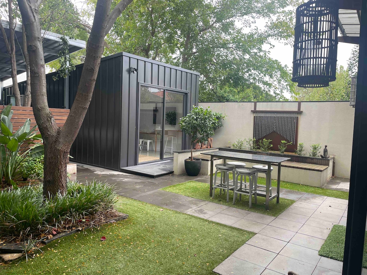A modern studio apartment is visible behind a well-maintained garden. Outdoor dining furniture is situated on a tiled patio, surrounded by lush greenery and potted plants. Large sliding glass doors connect the interior to the outdoor space, enhancing the overall light and openness.