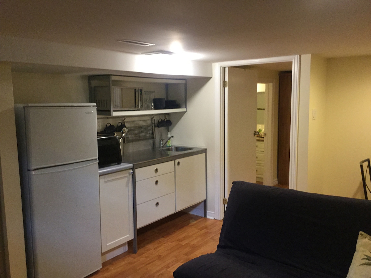 A compact kitchenette features a refrigerator, a small countertop space, and essential appliances, including a microwave and toaster oven. The cabinetry provides storage, while the adjacent door leads to the bedroom and bathroom. Natural light illuminates the warm-toned wooden flooring.