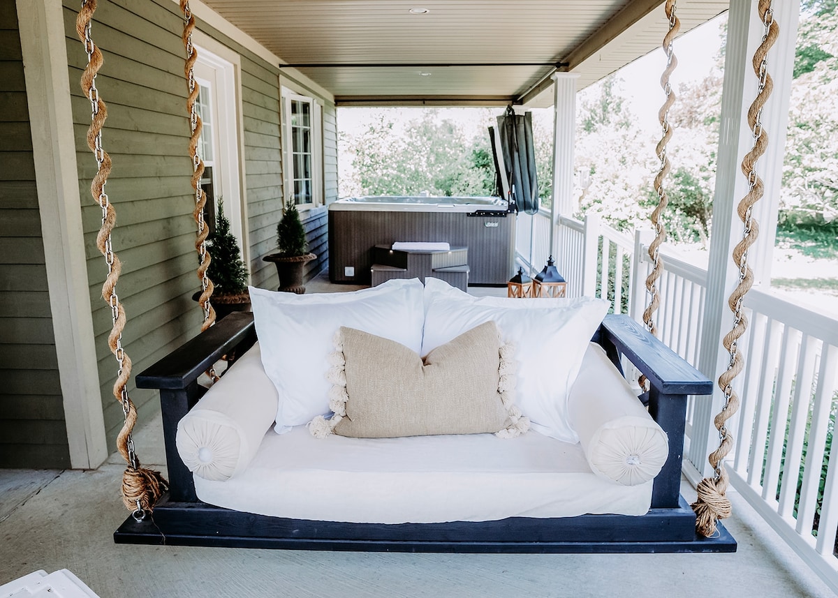 A spacious swing adorned with a neutral cushion and decorative pillows is positioned on a covered porch. The area features a jacuzzi in the background, surrounded by greenery, creating a relaxing outdoor space suitable for leisurely moments.