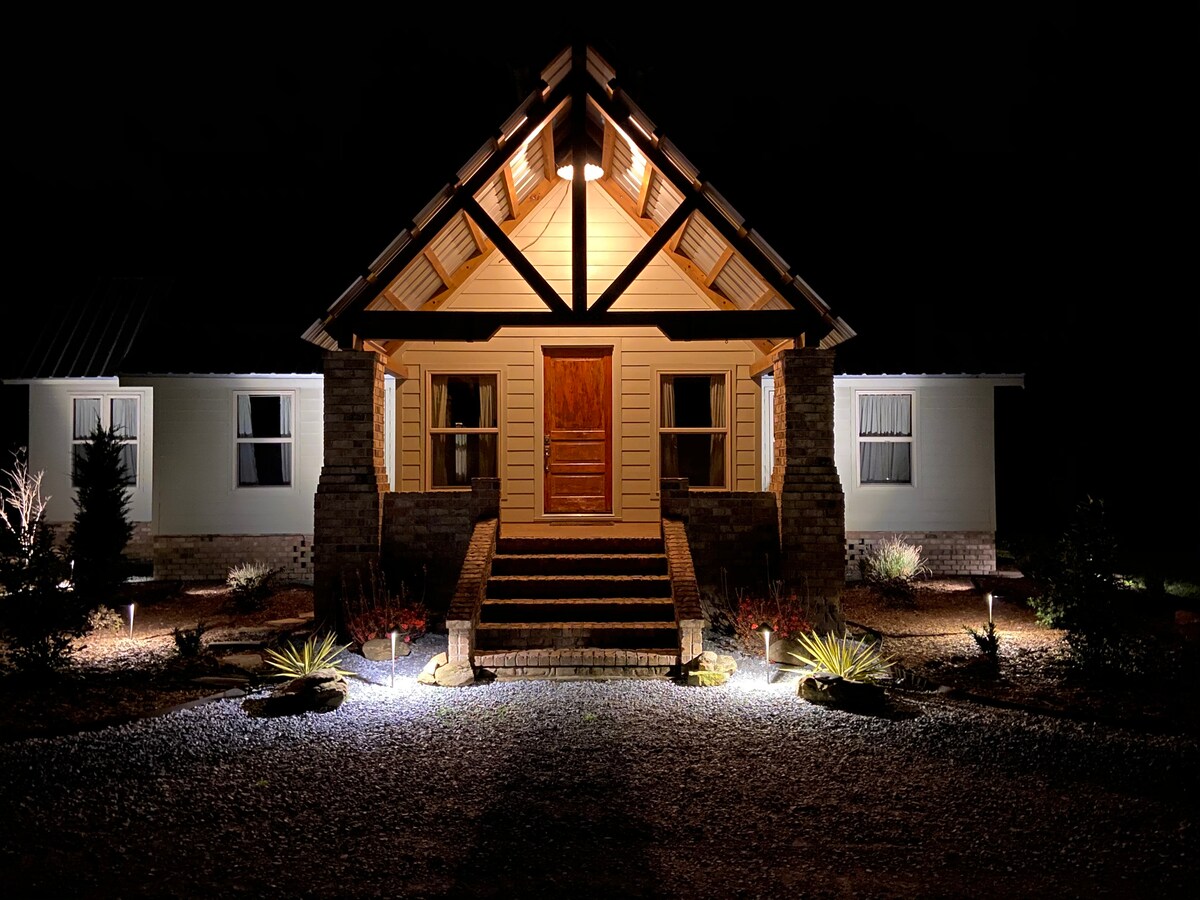 The exterior of a timber frame home is illuminated at night, showcasing its unique architectural design. Soft lighting highlights the stone steps leading to the wooden front door, flanked by landscaped areas featuring subtle greenery. The surrounding gravel driveway creates a welcoming approach.