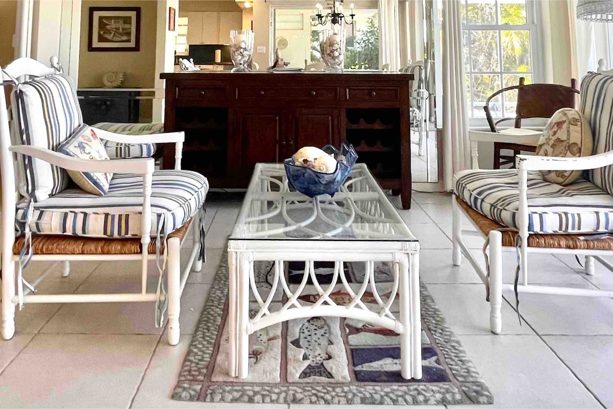 A welcoming living area features a glass-top coffee table surrounded by two armchairs with striped cushions and a sofa. Natural light floods the space through large windows, complementing the light-coloured tile flooring. A wooden sideboard with wine storage is visible in the background.