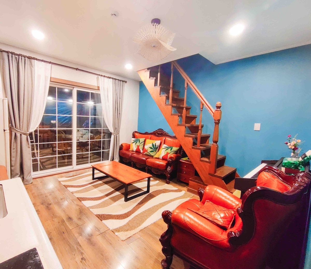 A living area is showcased, featuring a stylish staircase leading to an upper level. Two comfortable, red leather chairs flank a wooden coffee table on a light-patterned rug. Large windows allow natural light to fill the space, offering views of the outdoors.