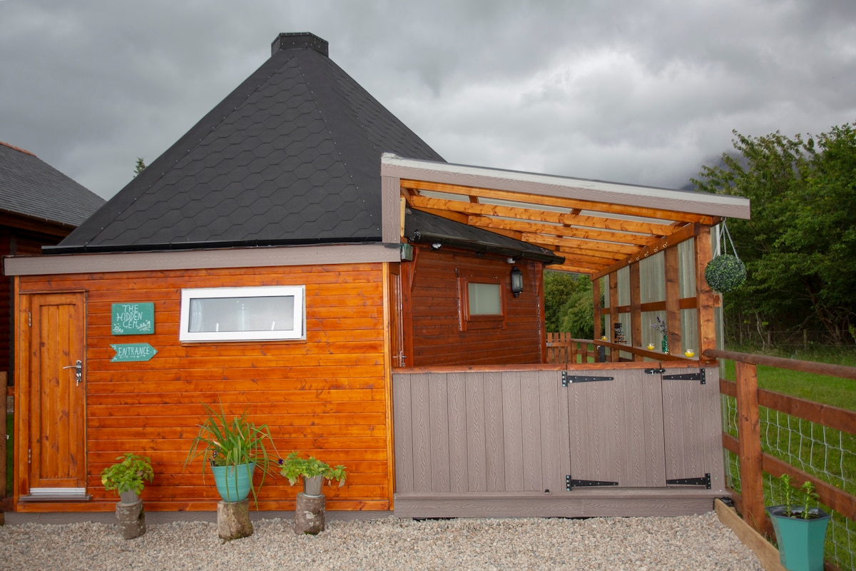 A chalet with a distinctive octagonal shape is presented, featuring warm wooden siding and a grey shingle roof. The entrance is highlighted by a wooden door and a decorative sign on the wall. A sheltered decking area is visible, surrounded by planters with greenery.