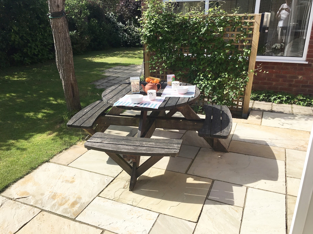An outdoor picnic table is set with plates, cups, and a selection of food items. The surrounding area features well-maintained paved stones and a manicured lawn. Green shrubbery provides a natural backdrop, while large windows of the flat allow for natural light.