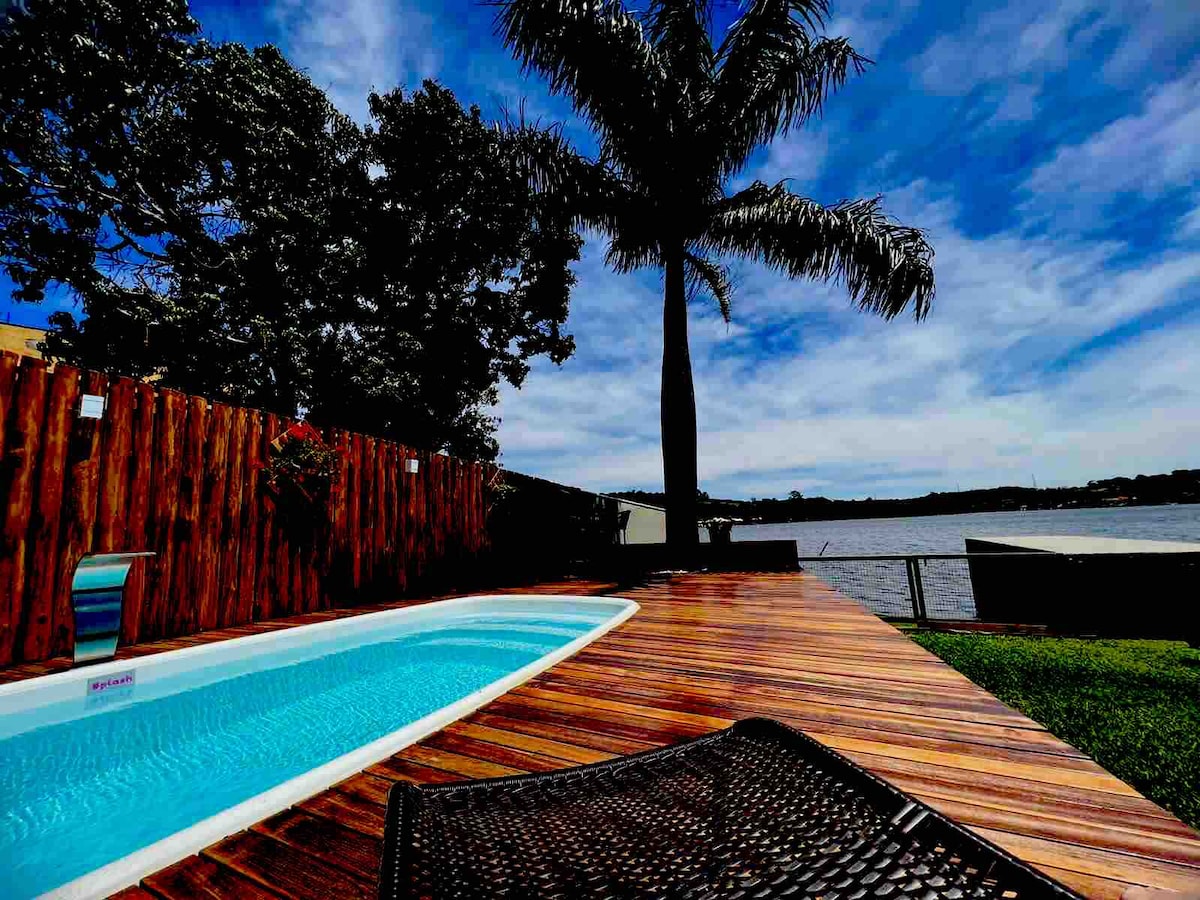 A view of a modern swimming pool surrounded by wooden decking, with lush green grass nearby. A tall palm tree stands elegantly against a vibrant blue sky, providing a tropical feel. The serene waters of the reservoir are visible in the background.