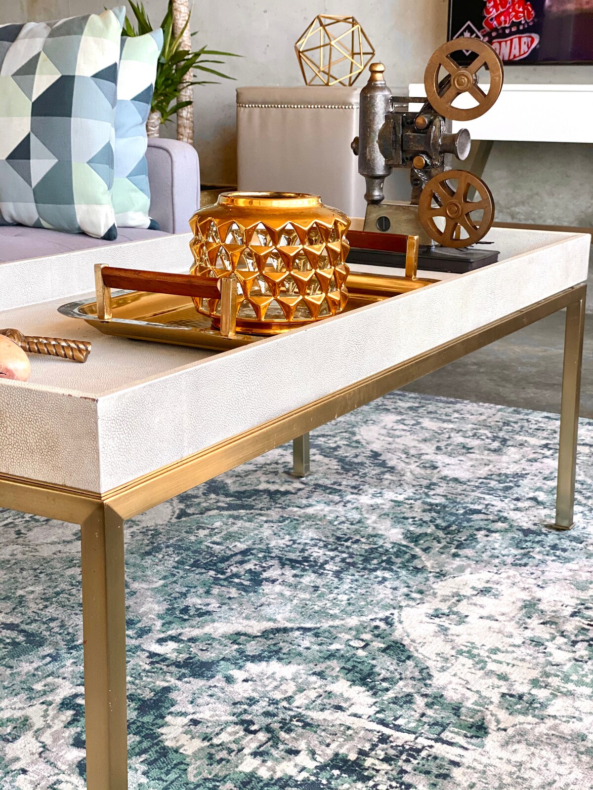 A stylish coffee table is centered in a living area, featuring a light-colored textured surface and gold metal legs. An ornate golden candle holder, a vintage decorative piece, and a tray are arranged on top, while a patterned area rug adds a soft touch underneath.