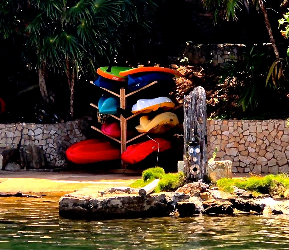 A stack of kayaks in various colors sits on a stand near the water's edge, surrounded by lush greenery. The calm surface of the water reflects the vibrant hues, while a stone structure is positioned nearby, enhancing the natural setting.