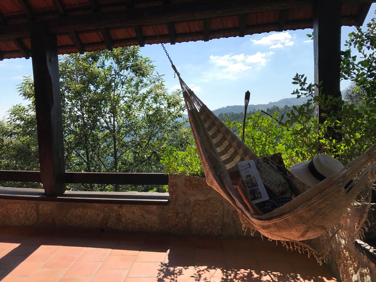A hammock is gently swaying in a spacious covered terrace, offering views of lush greenery and distant hills. A cozy blanket and a straw hat are nestled within the hammock. Natural light filters through the open space, creating a serene atmosphere.