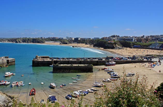 A scenic view of a beach and harbor in Newquay, with boats anchored along the shoreline. The sandy beach is dotted with people enjoying the sun, while cliffs and coastal buildings frame the background. The vibrant blue sea reflects the clear sky overhead.
