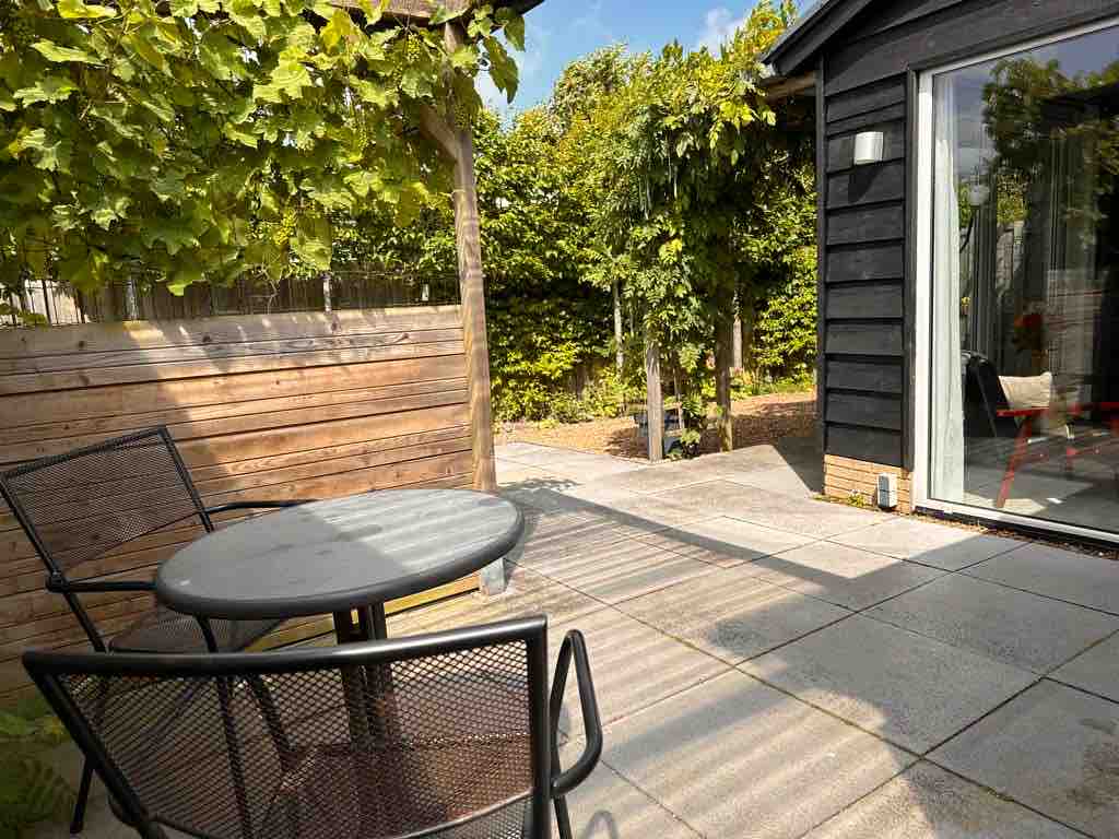 A shaded outdoor space features a round table and two chairs set upon a tiled patio, surrounded by lush greenery. The space offers a serene setting, with sunlight filtering through the leaves providing a calming ambiance.
