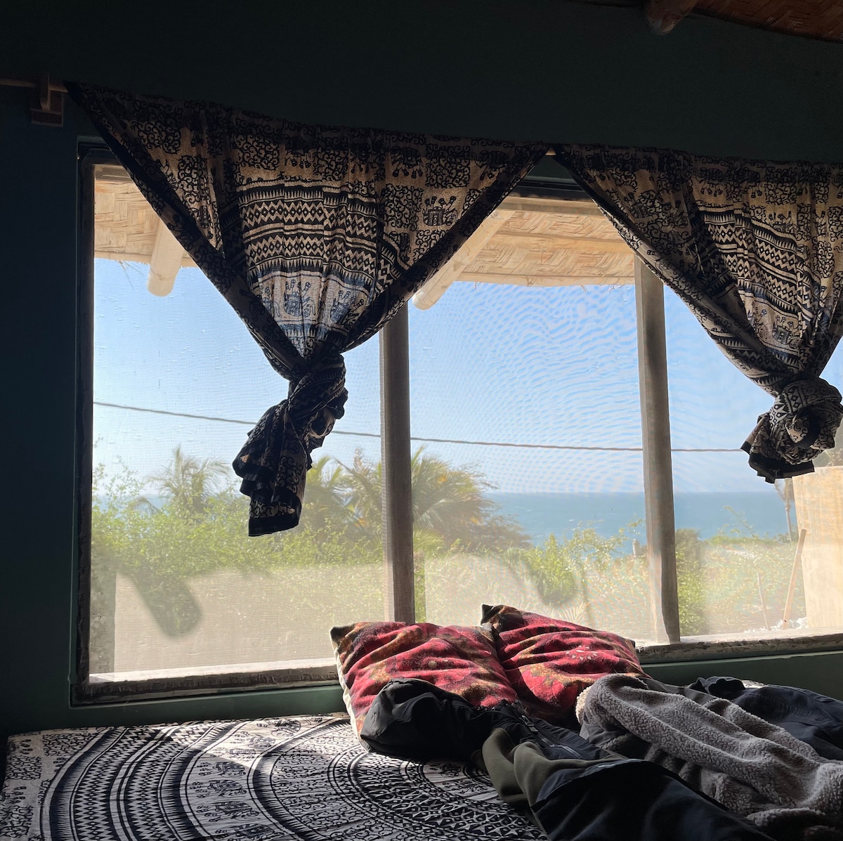 A window with lace curtains frames a scenic view of the beach. The view showcases the ocean and lush greenery. On the bed, colorful blankets are laid out, creating a cozy atmosphere within the room.