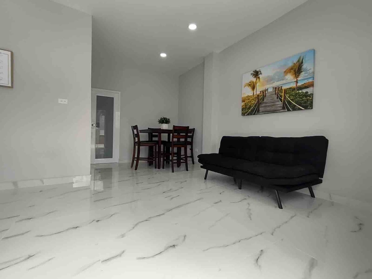 An open living space is highlighted by glossy marble floors and neutral-colored walls. A sleek black sofa faces a dining area furnished with a wooden table and four chairs. A large wall art piece depicting a beach scene adds a touch of color to the room.