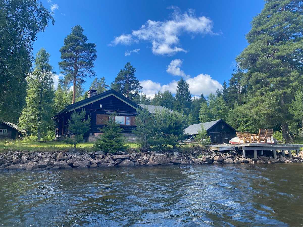 A charming wooden cabin is situated by the water, framed by lush greenery. The exterior features large windows and a spacious deck with seating. Surrounding trees create a natural border, enhancing the serene lakeside setting under a bright blue sky with scattered clouds.
