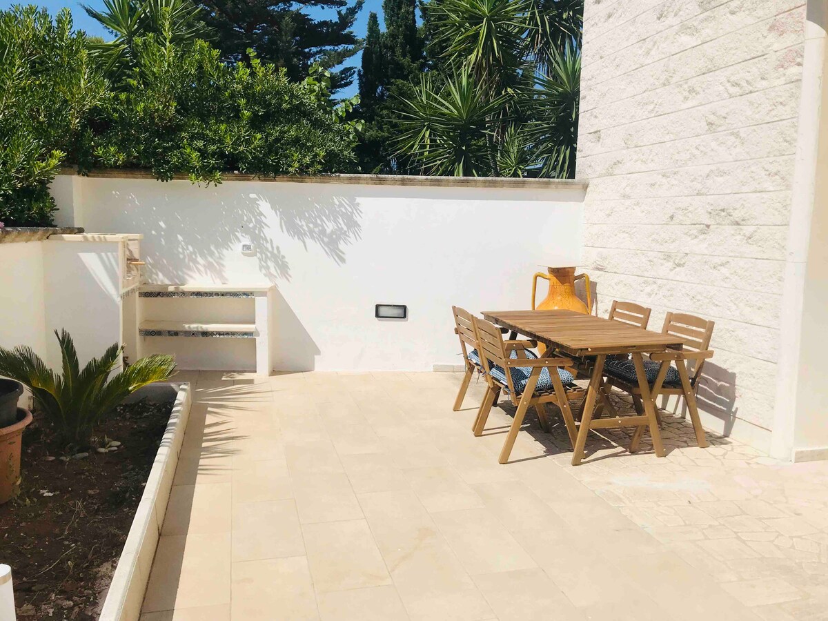 A private outdoor space is presented, featuring a wooden dining table surrounded by six chairs. Lush greenery is visible in the background, while a small barbecue corner is positioned against a white wall. The floor is tiled with light-colored stone.