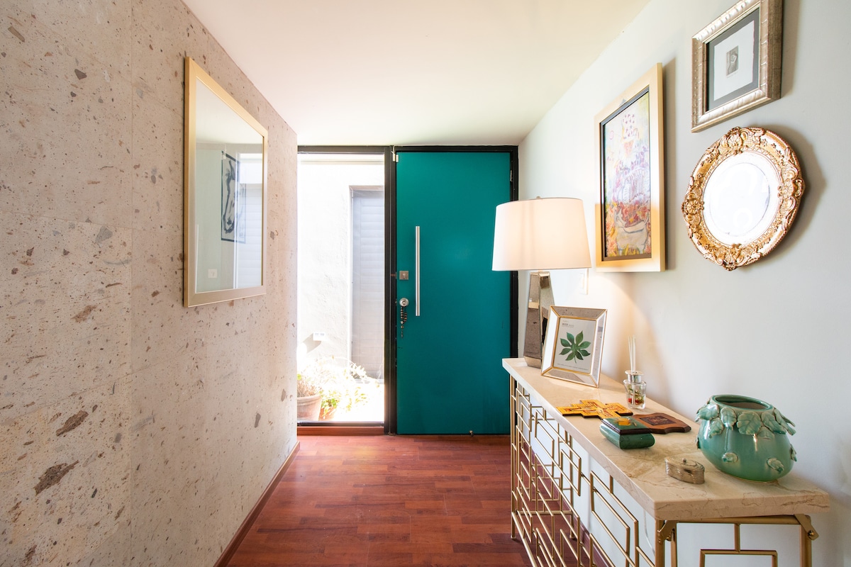 A welcoming entrance features a vibrant teal door and a stylish console table. Framed artwork and a decorative plate adorn the walls, while natural light enters from a glass-panel door, highlighting the textured wall and wooden flooring.