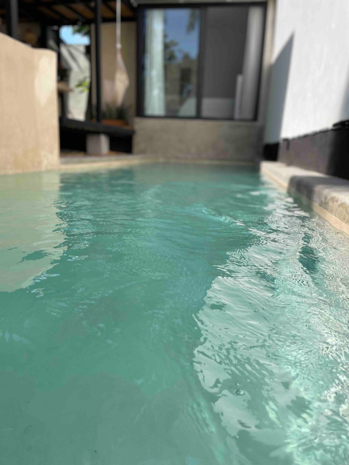 A turquoise swimming channel glistens in the sunlight, reflecting the surroundings. The smooth concrete edges create a clean and modern design, while a glimpse of large glass doors and a tranquil indoor space is seen in the background.