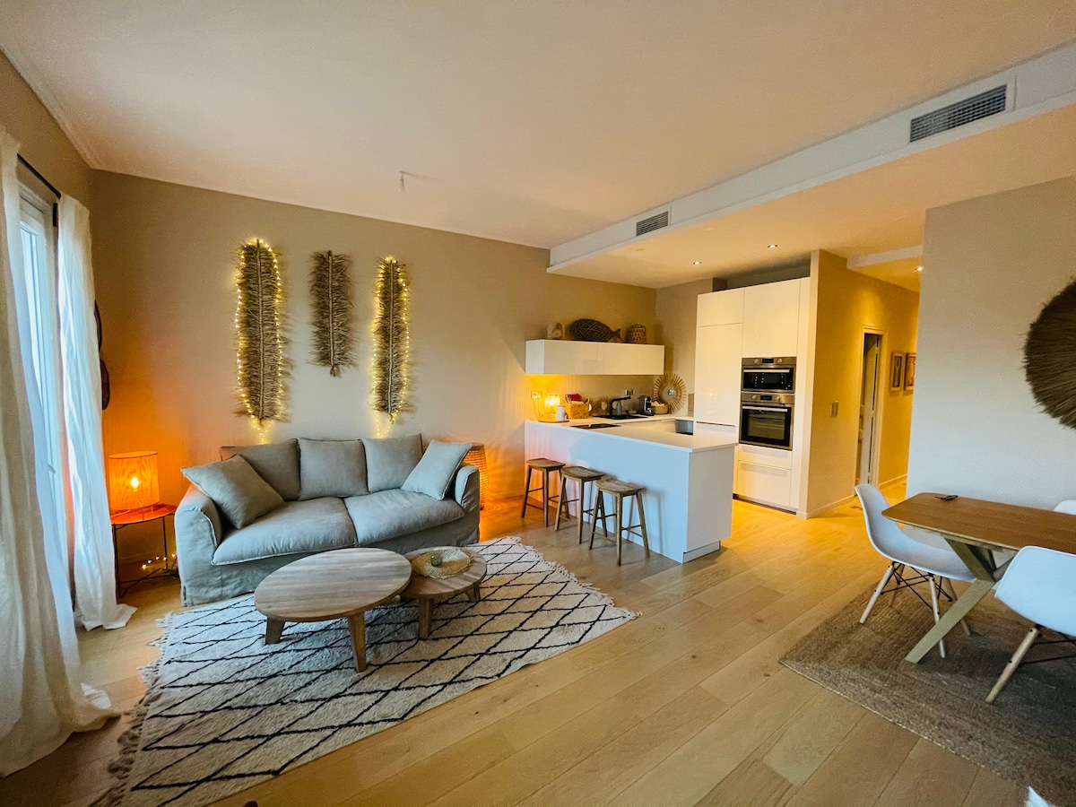 A modern living space features a comfortable sofa, light wood flooring, and a round wooden coffee table. The kitchen is equipped with sleek appliances, and a dining area with white chairs is visible. Soft lighting enhances the welcoming atmosphere.