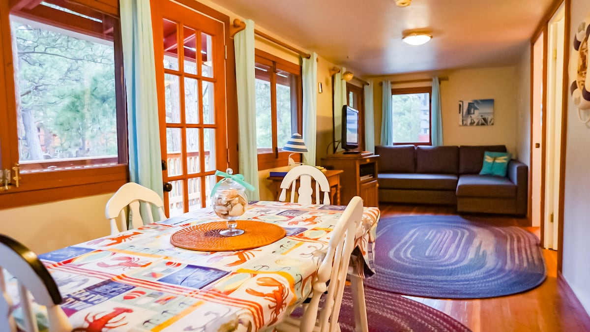 A dining area features a table with a colorful tablecloth, set for four. Natural light fills the space through large windows, highlighting a cozy seating area with a gray couch. The warm wood tones of the furniture provide a welcoming ambiance.