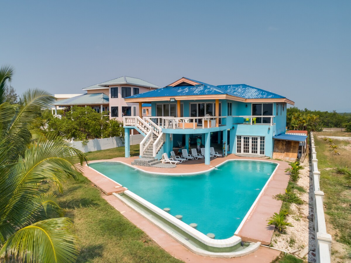 An oceanfront villa showcases vibrant blue exterior walls with a contrasting orange roof. The infinity pool is surrounded by a spacious deck with lounge chairs, and a staircase leads to a second-level entrance. Lush greenery frames the property, enhancing the serene outdoor setting.