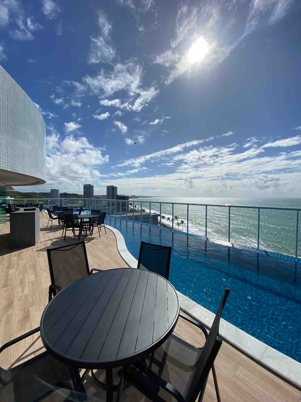 A panoramic view of the ocean unfolds from a rooftop terrace, featuring several black outdoor tables and chairs. A clear blue infinity pool extends towards the horizon, with a partly cloudy sky overhead, brightened by the sun's reflection on the water.