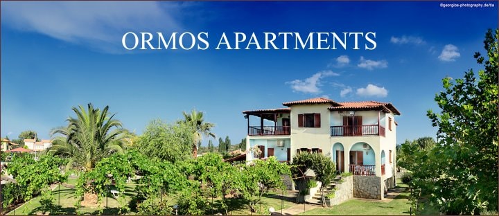 The image captures the exterior of Ormos Apartments, a two-story building surrounded by lush greenery and palm trees. Balconies are visible on each floor, providing outdoor spaces for relaxation. A clear blue sky enhances the inviting setting, highlighting the peaceful environment.