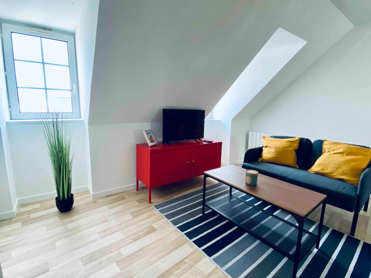 A small living area is illuminated by natural light from a nearby window. A dark sofa adorned with yellow cushions faces a low coffee table. A red cabinet holds a television, while a small plant adds a touch of greenery to the space.