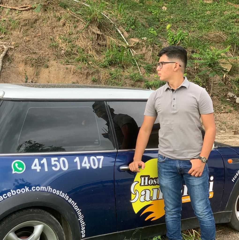 A compact car is parked beside a grassy area, displaying a colorful logo on its side. A person is seen standing next to the vehicle, wearing a gray polo shirt and blue jeans. The background features a lightly wooded hillside.