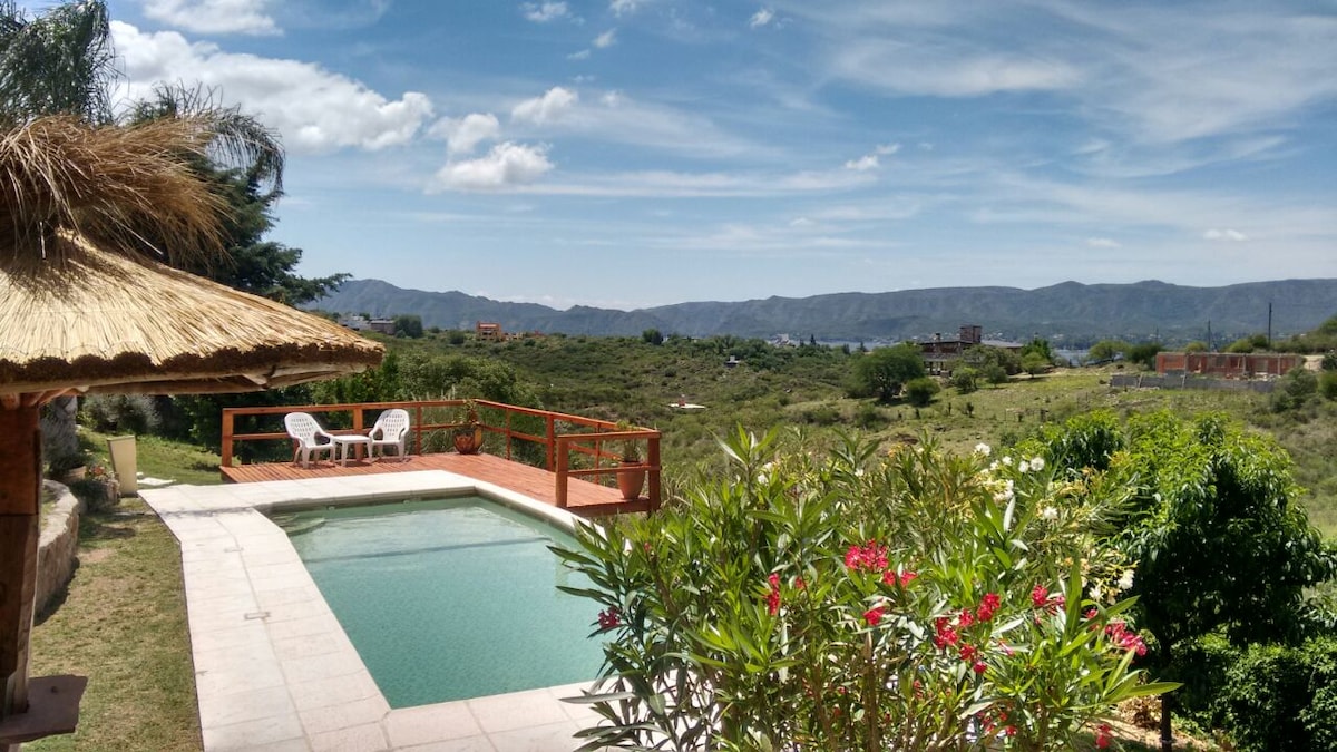 A private pool is surrounded by lush greenery and positioned beside a wooden deck. Two white chairs are placed on the deck, offering a view of the mountains and valleys in the distance under a bright blue sky with scattered clouds.