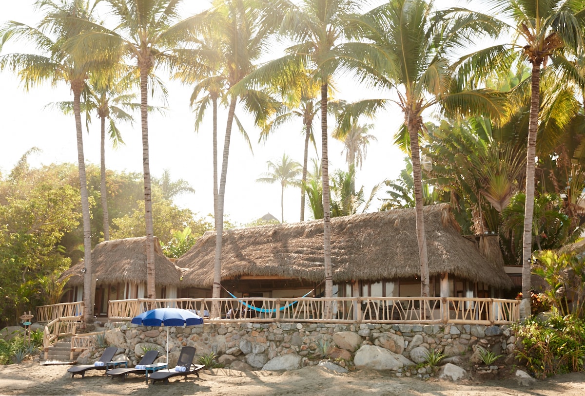 A beachfront bungalow is surrounded by tall palm trees and lush greenery, featuring a thatched roof and a stone wall. Outdoor seating and a hammock are visible on the terrace, with a blue umbrella and lounge chairs placed on the sand for relaxation.