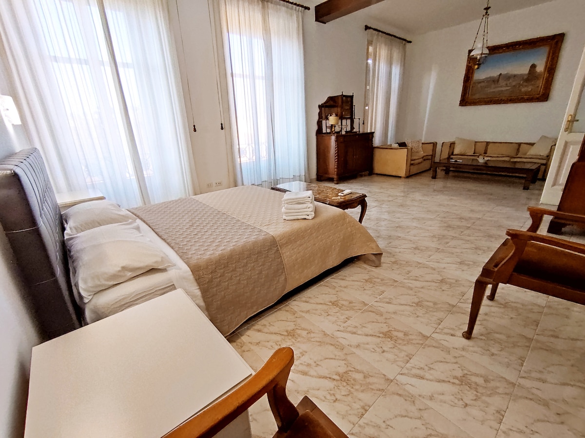 A spacious bedroom features a large double bed with a neutral quilt, complemented by two soft pillows. Floor-to-ceiling windows allow natural light to fill the room, while elegant drapes add a touch of sophistication. A comfortable sitting area is arranged with wooden chairs and a glass coffee table.