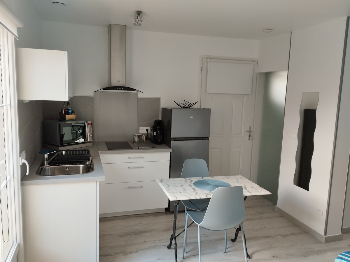 A modern kitchen area features sleek white cabinetry and stainless steel appliances. A compact dining table with two grey chairs is positioned near a large window, allowing natural light to fill the space. Counter space is utilized for food preparation and coffee making.