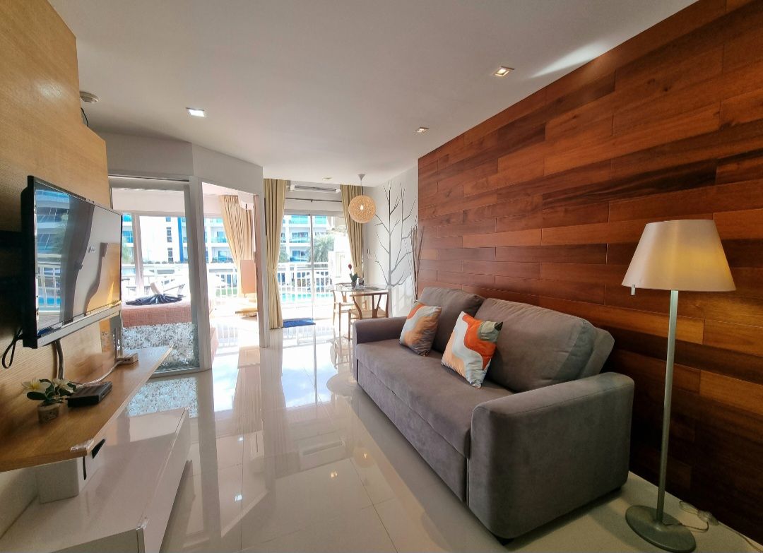 A living area features a plush gray sofa adorned with accent pillows, positioned next to a modern lamp. A wooden accent wall adds warmth, while large windows provide natural light and access to outdoor views. A dining table is visible in the background.